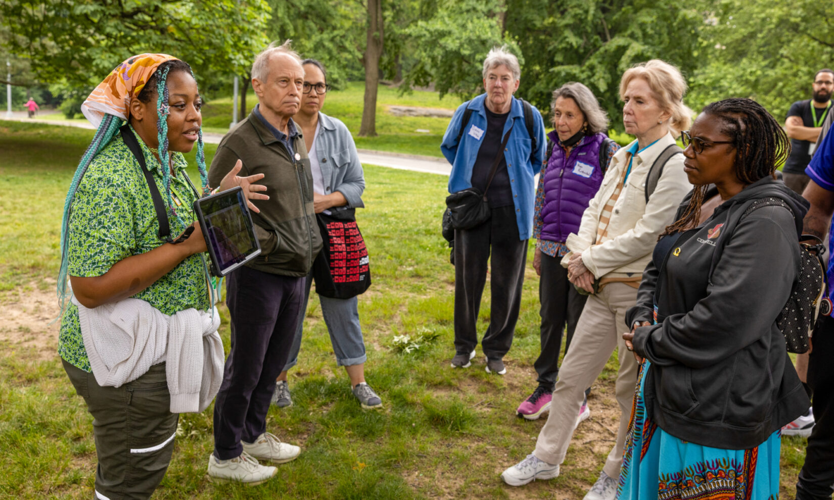 A group of people stand in a grassy area and listen to a person talking and showing them a tablet.