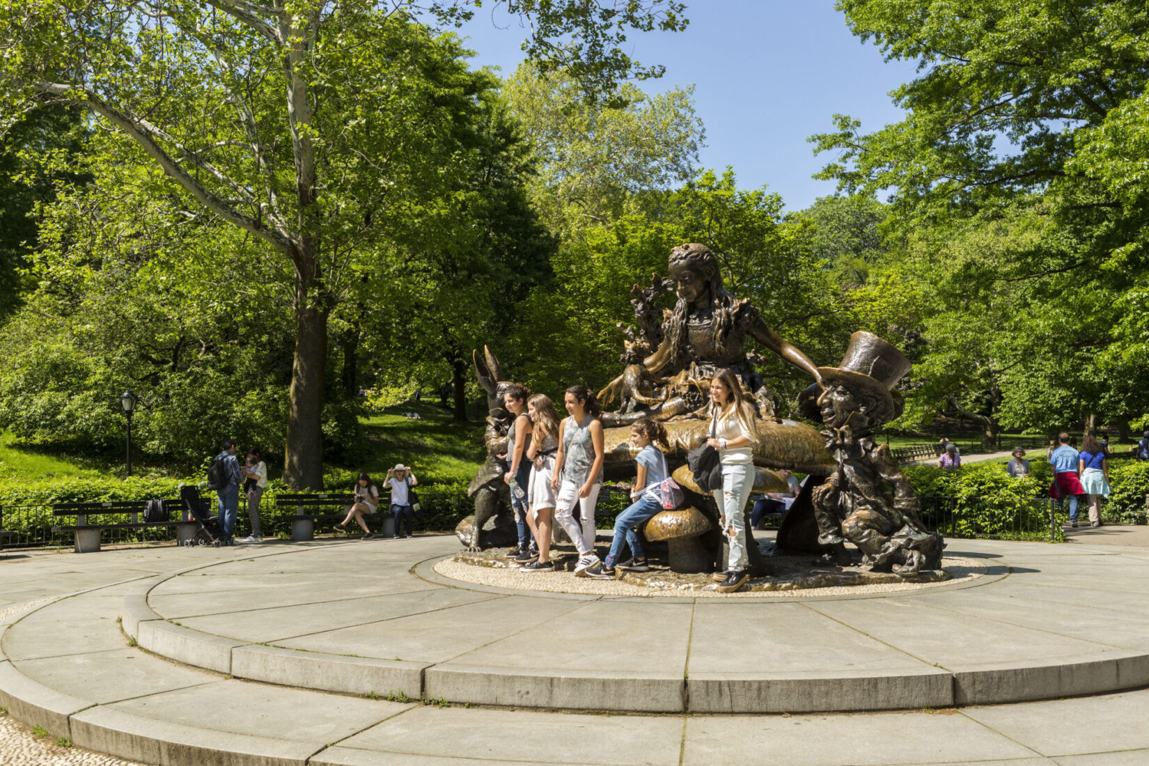 A group of children and an adult smile while standing in front of a statue of characters from Alice in Wonderland.