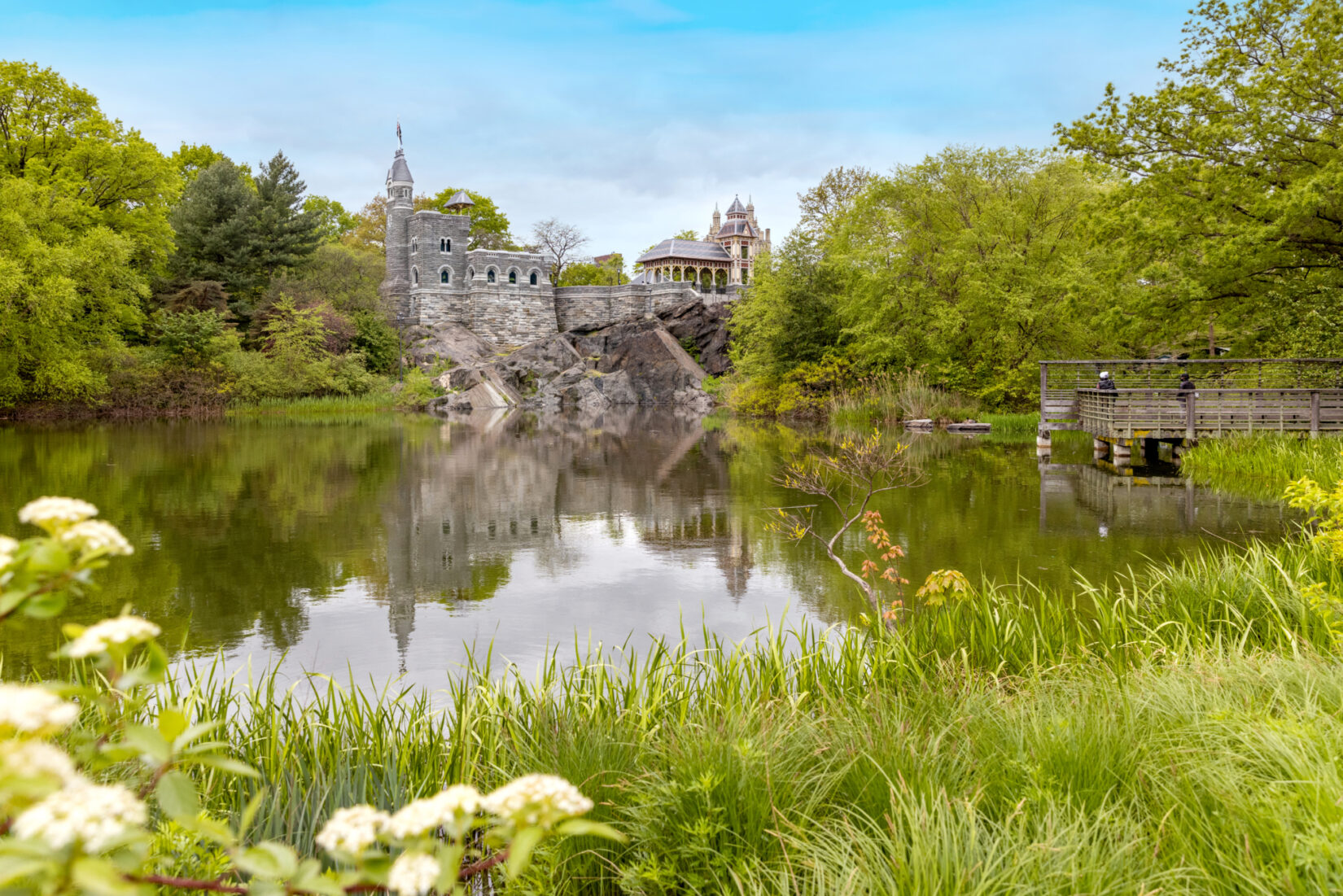 A grey, stone castle stands behind a small body of water, surrounded by bright green trees and grass.