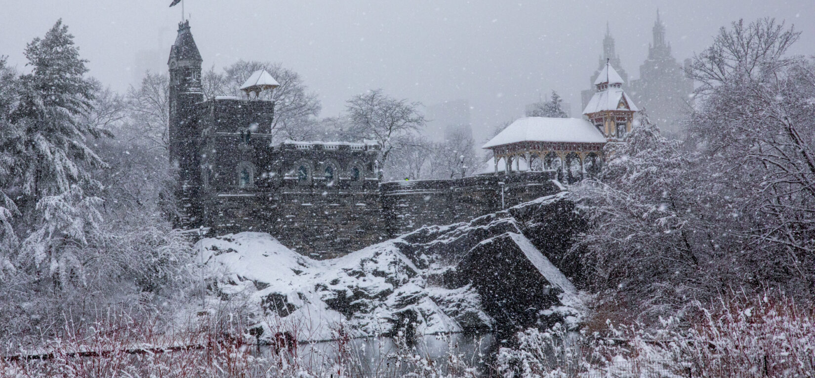 Belvedere castle surrounded by rocks and trees covered with snow, and a small body of water in front of it.