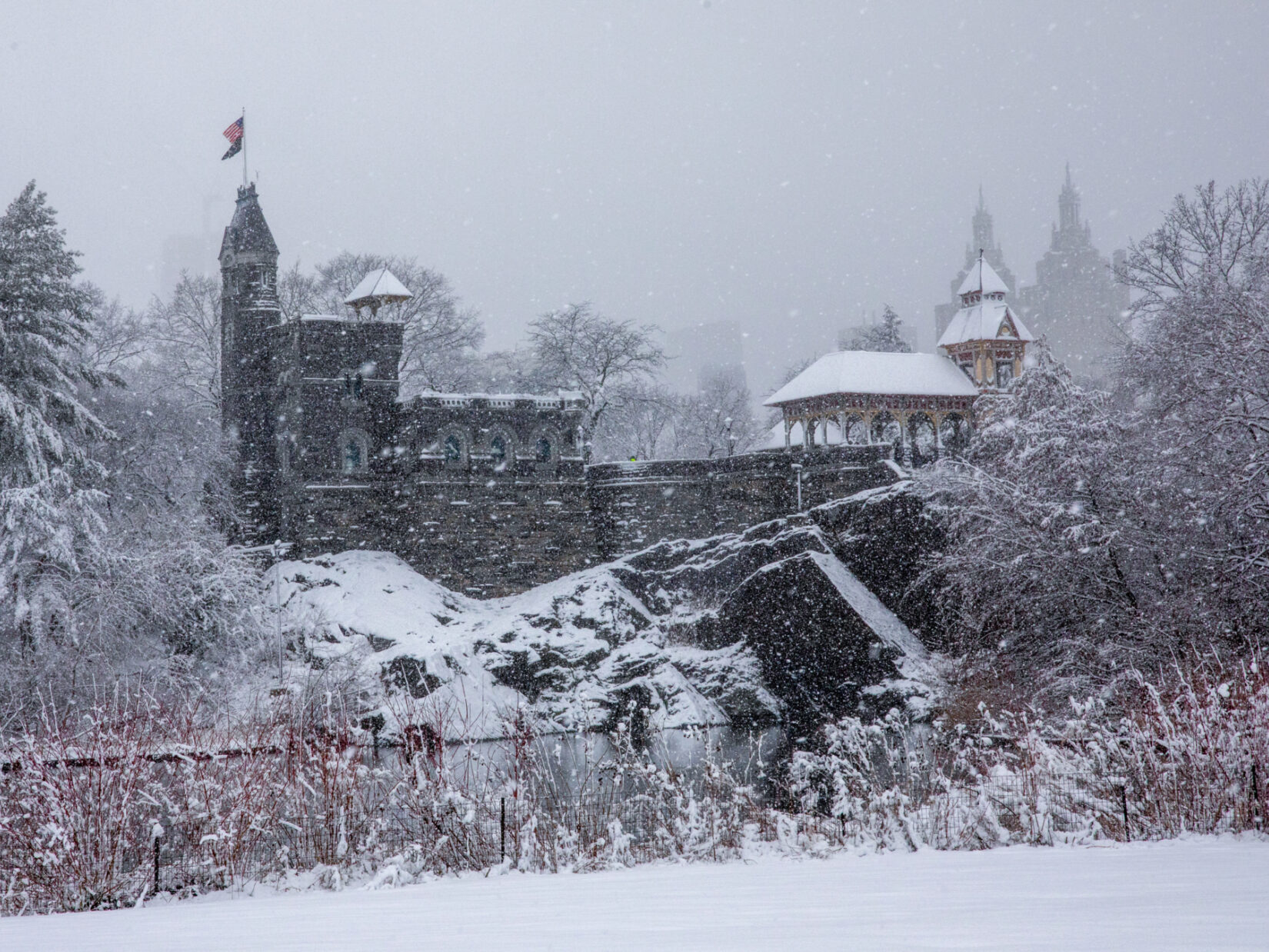 Belvedere castle surrounded by rocks and trees covered with snow, and a small body of water in front of it.