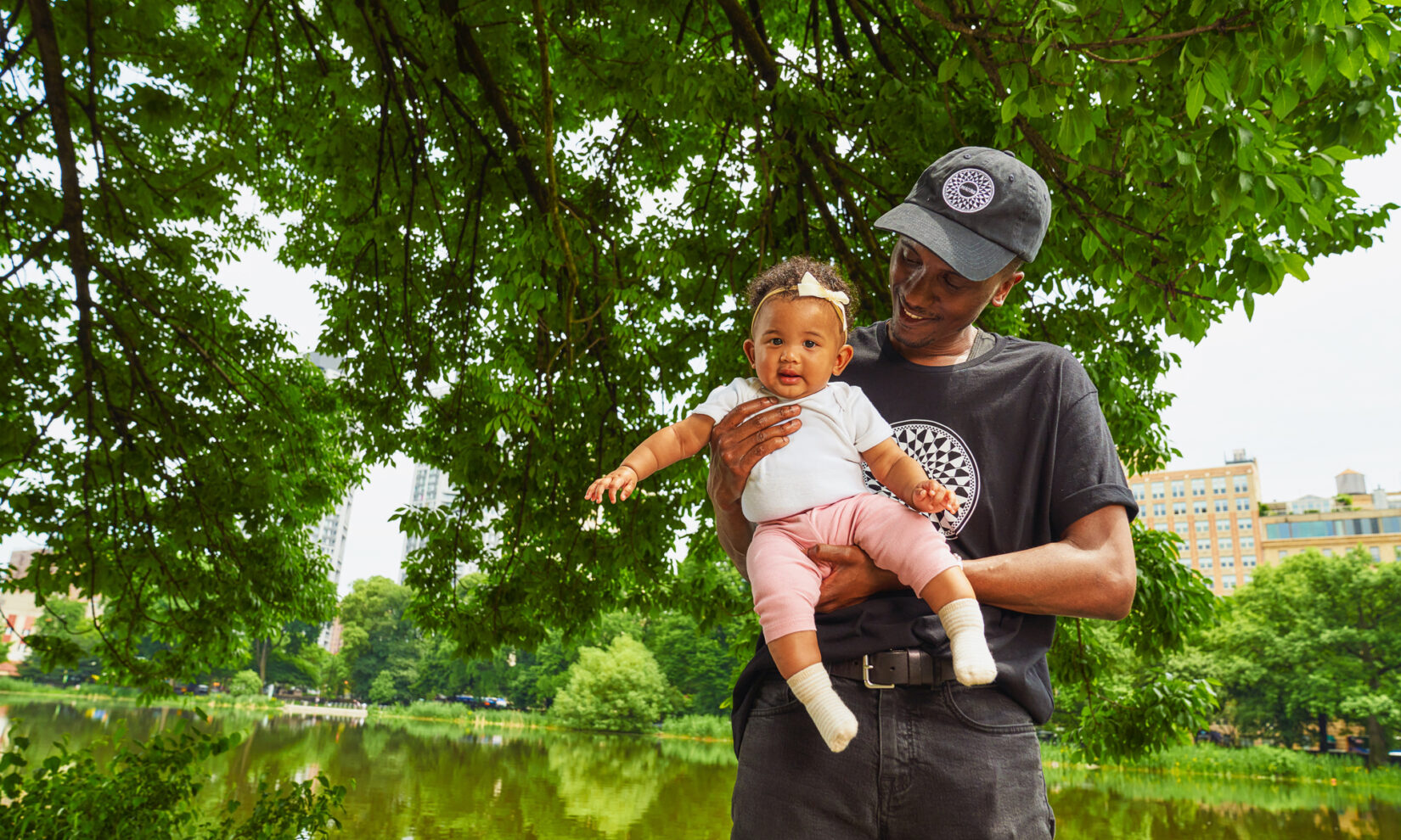 A man in a Central Park t-shirt holds a baby in a Park onesie, with trees and pond in background